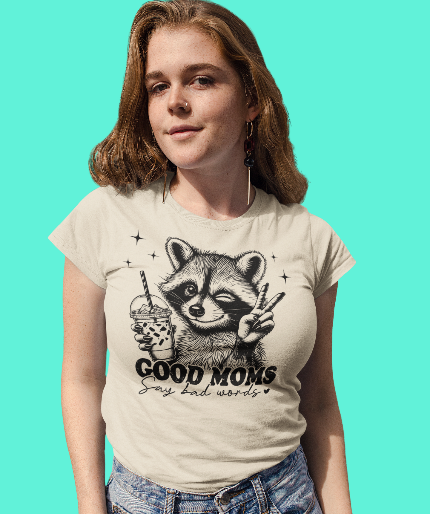 Γυναίκα με μπεζ μπλουζάκι που γράφει "Good Moms Say Bad Words" με ρακούν, ποζάρει μπροστά σε γαλάζιο φόντο με χαλαρό χαμόγελο.