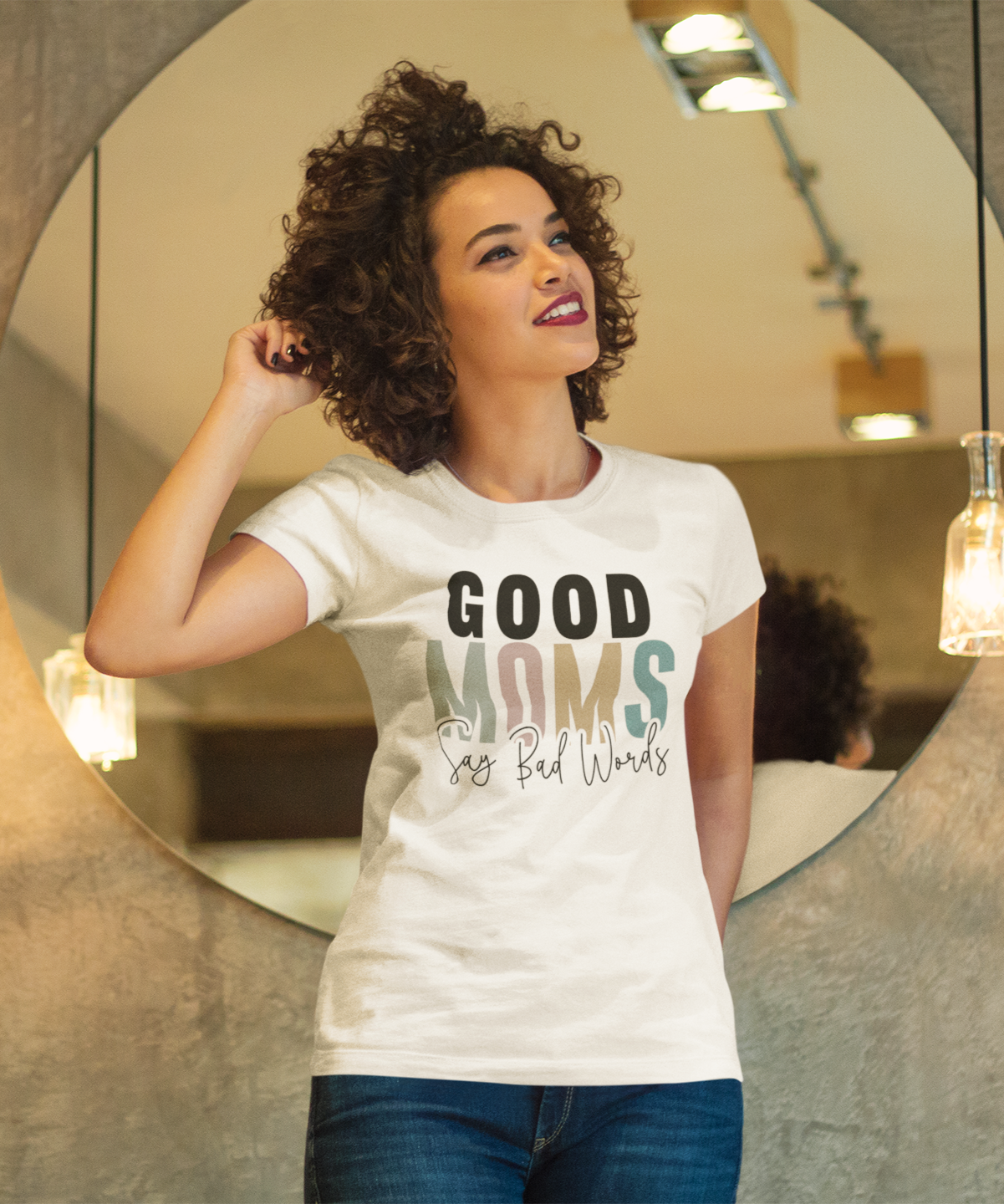 μπλουζάκι που γράφει "Good Moms Say Bad Words", στέκεται μπροστά από καθρέφτη σε μοντέρνο χώρο με φωτιστικά.